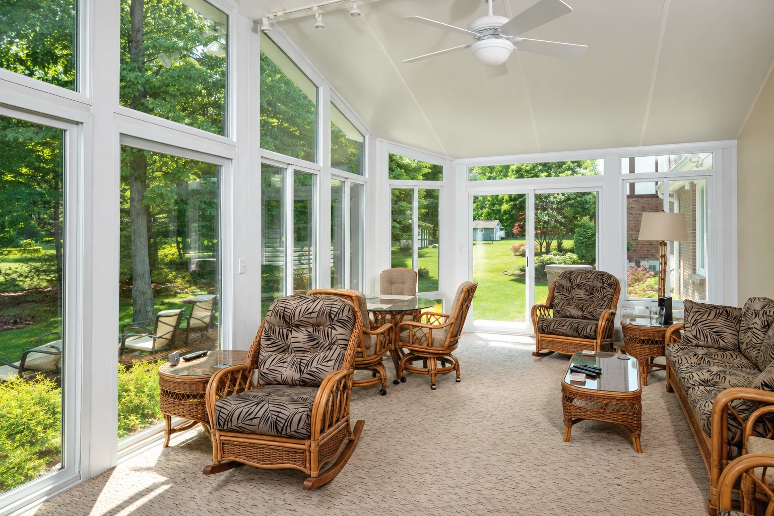 Bright four-season sunroom with wicker seating, a ceiling fan, and tall windows overlooking a leafy garden.