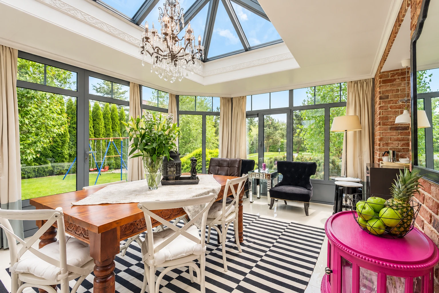 Four-season sunroom with a glass-paneled peaked ceiling, dining table, and dark-framed windows overlooking a garden.