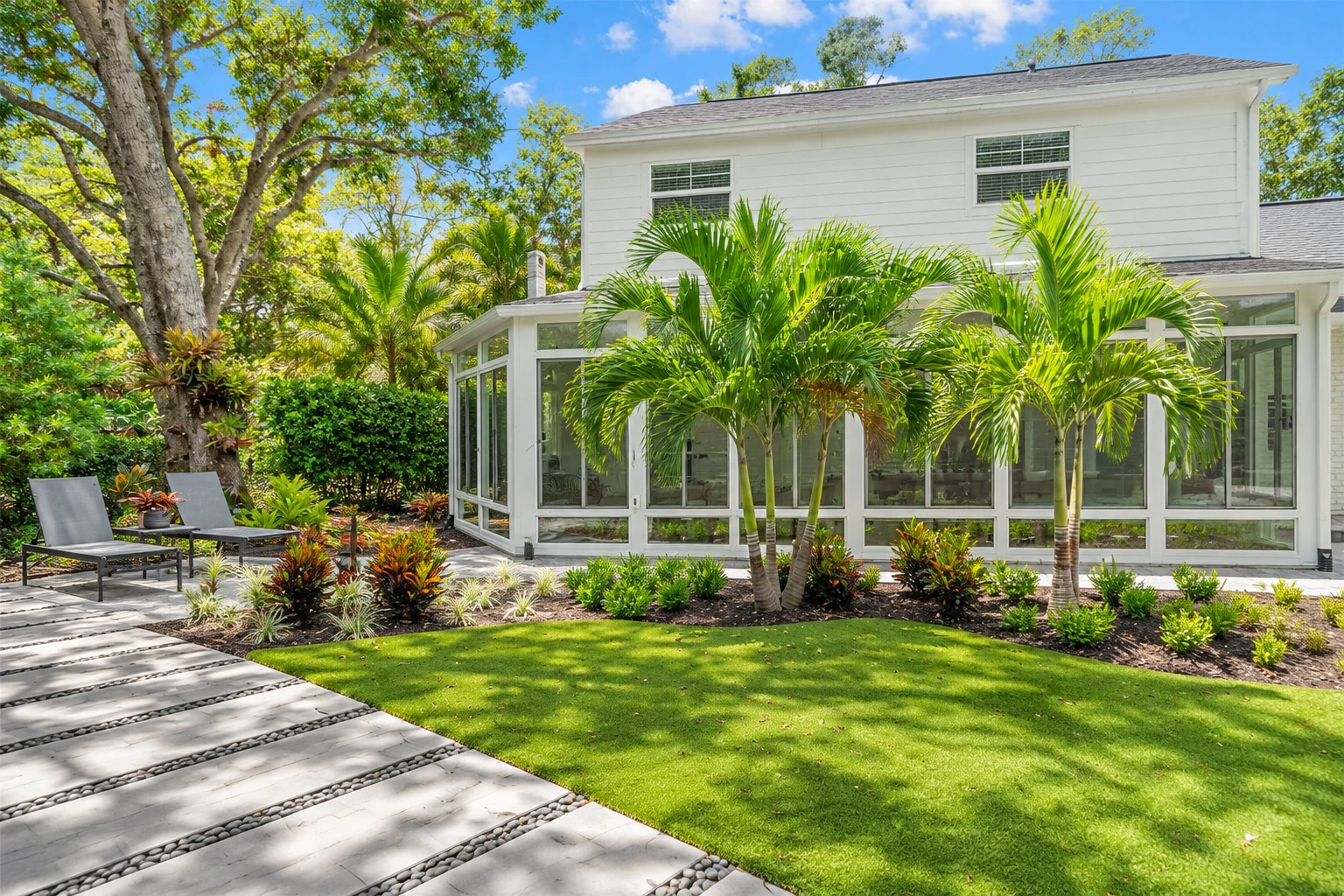 White-framed glass patio enclosure beside a house, surrounded by palms, tropical plants, and a lawn.