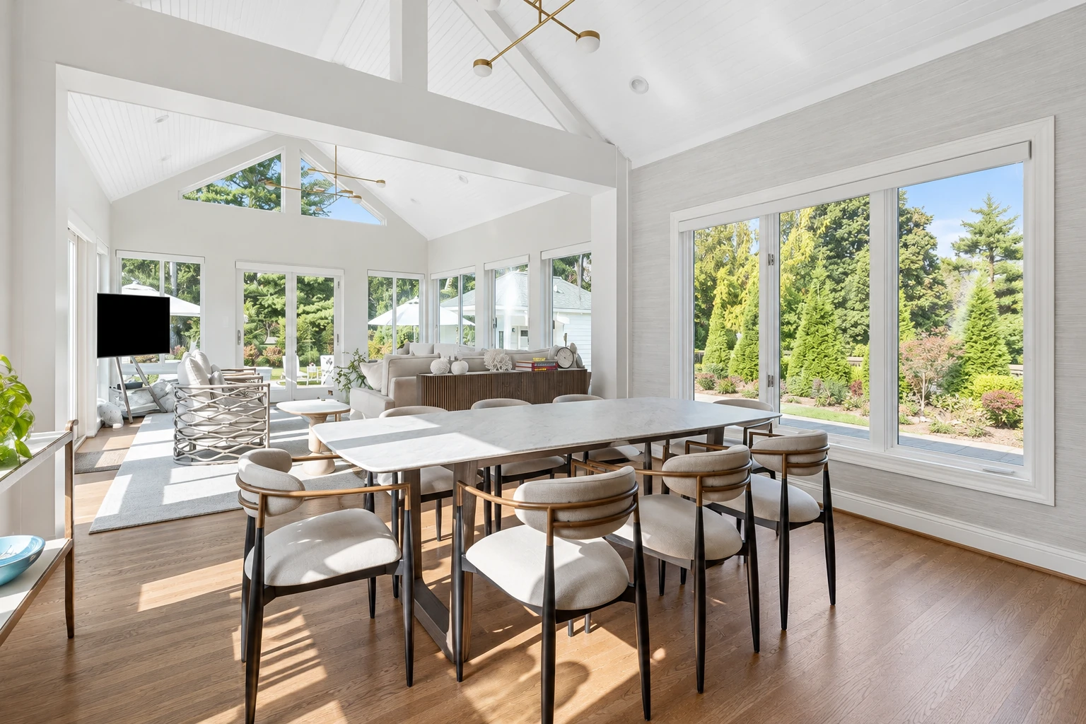 Bright sunroom with a high white vaulted ceiling, a long dining table, and large windows overlooking a garden.