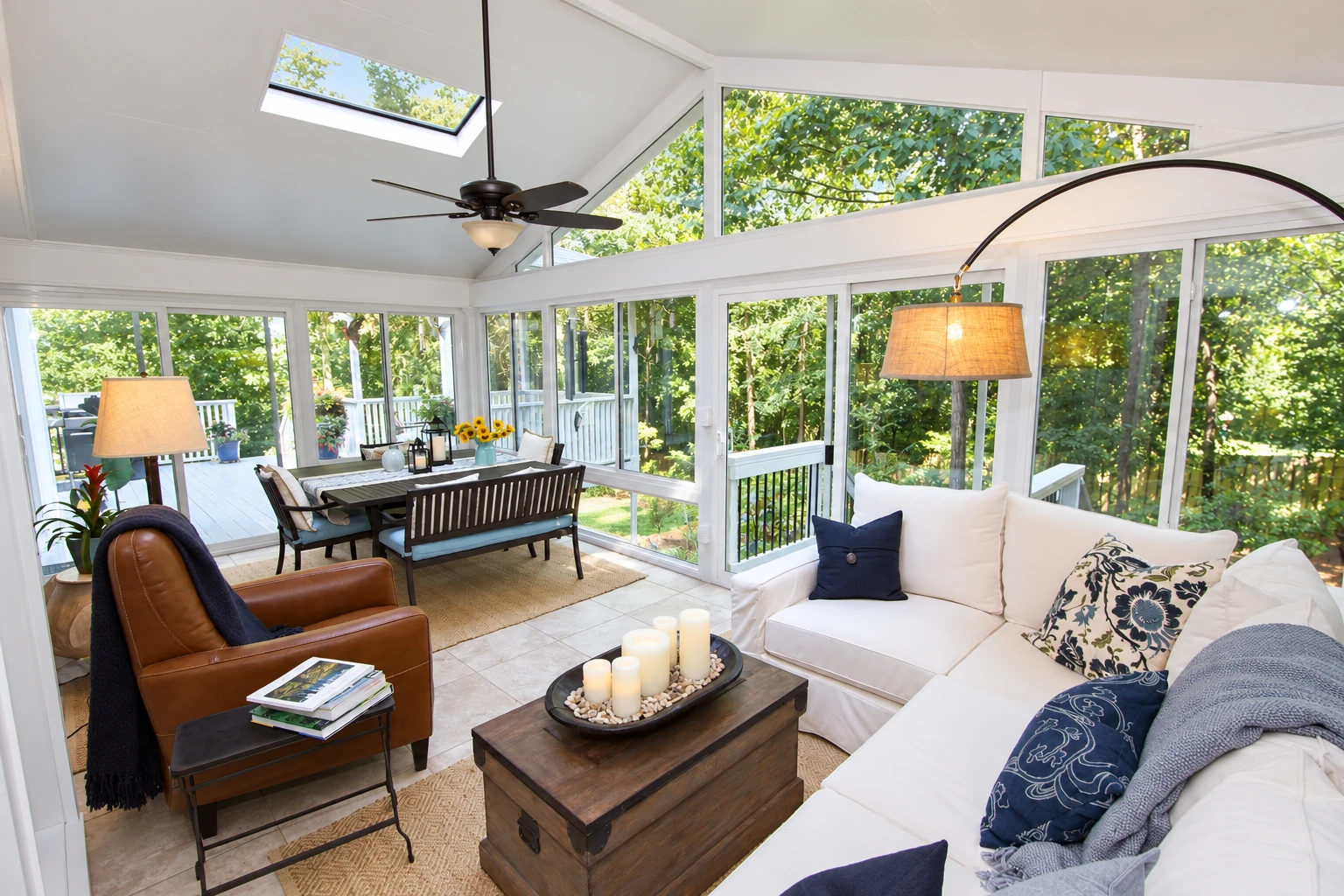 Bright sunroom with white-framed windows, skylight, dining area, and seating overlooking leafy trees.